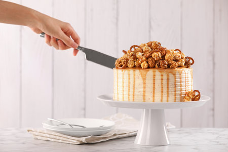 Woman cutting caramel drip cake decorated with popcorn and pretzels at white marble table, closeup