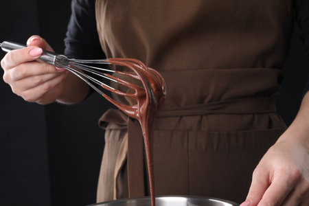 Woman holding whisk with flowing chocolate cream on black background, closeup
