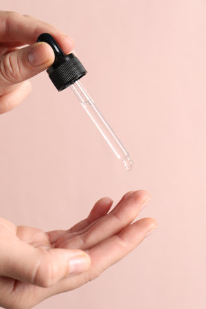 Woman applying cosmetic serum onto fingers on light pink background, closeup