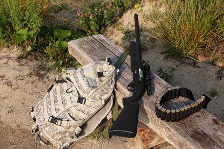 Hunting rifle, backpack and cartridges on wooden bench outdoors