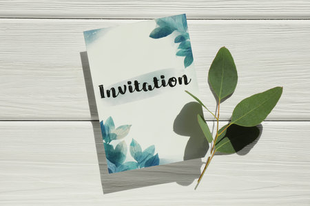Beautiful card with word Invitation and green leaves on white wooden table, top view