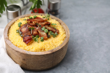 Cooked cornmeal with bacon and microgreens in bowl on light gray table, closeup. Space for text