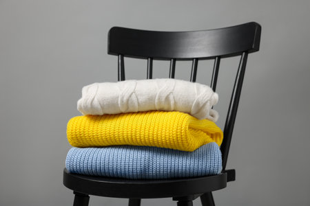 Stack of different casual sweaters on black chair against gray background