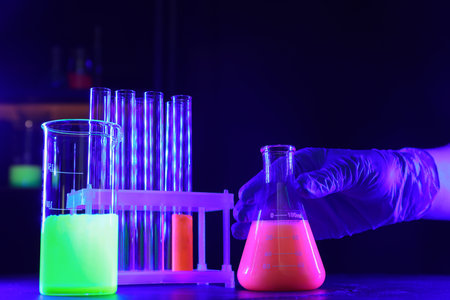 Scientist working with laboratory glassware of luminous liquids at table against dark background,...