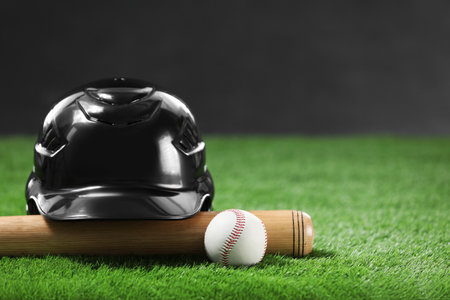 Baseball bat, batting helmet and leather ball on green grass against dark background, closeup. Space...