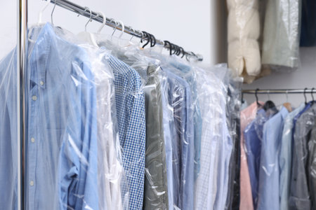 Dry-cleaning service. Hangers with different clothes in plastic bags on racks indoors, space for text
