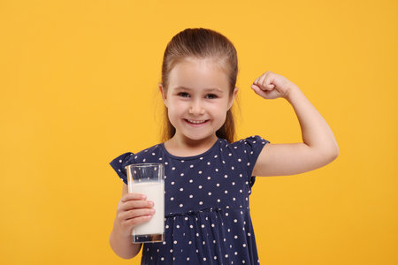 Cute girl with glass of fresh milk showing her strength on orange backgroundの写真素材