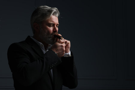 Bearded man lighting cigar on dark gray background. Space for text