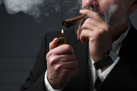 Bearded man lighting cigar on dark gray background, closeupの写真素材