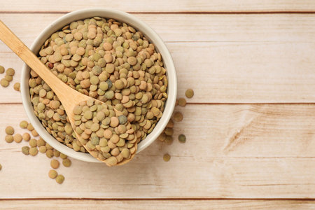 Raw lentils in bowl and spoon on light wooden table, top view. Space for text