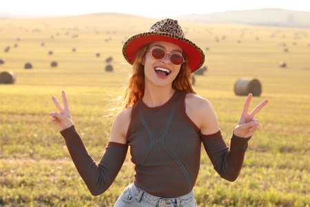 Beautiful happy hippie woman in hat showing peace signs in fieldの写真素材