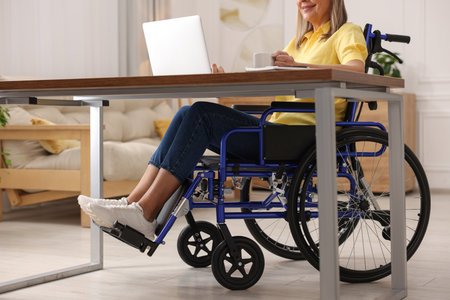 Woman in wheelchair with cup of drink using laptop at home, closeup