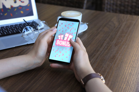 Bonus gaining. Woman using smartphone at wooden table, closeup. Illustration of open gift boxes,...