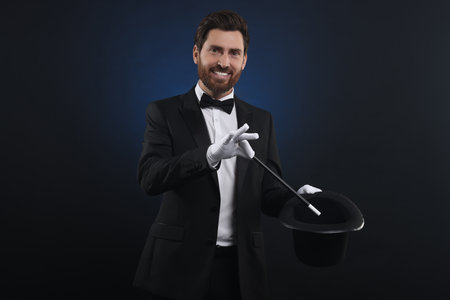 Happy magician showing magic trick with top hat on dark blue background