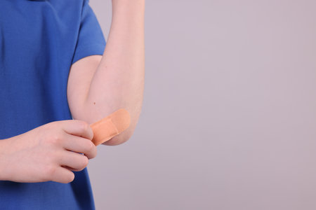 Little boy putting sticking plaster onto elbow against light gray background, closeup. Space for...