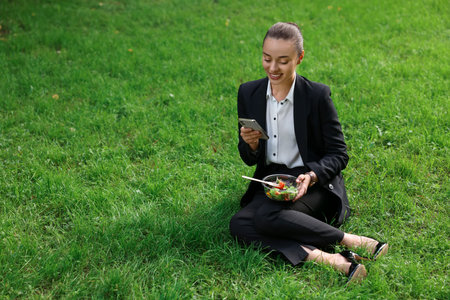 Lunch time. Happy businesswoman with container of salad using smartphone on green grass outdoors, space for textの写真素材