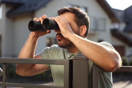 Concept of private life. Curious man with binoculars spying on neighbors over fence outdoorsの写真素材