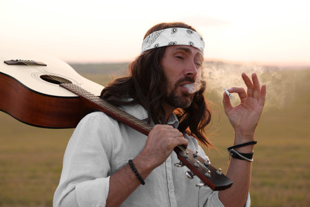 Stylish hippie man with guitar smoking joint in fieldの写真素材