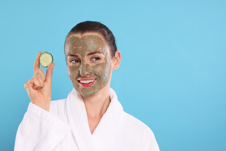 Beautiful woman with clay mask holding piece of cucumber on light blue background, space for textの写真素材