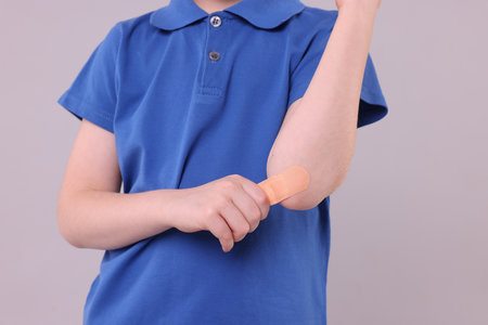 Little boy putting sticking plaster onto elbow against light gray background, closeup