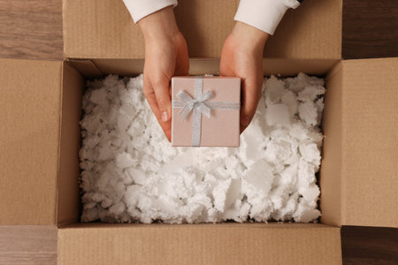 Woman putting Christmas gift box into parcel at wooden table, top view. Sending present by mail