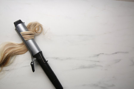 Curling iron with blonde hair lock on white marble background, top view. Space for textの写真素材
