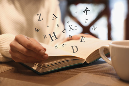 Woman with coffee reading book with letters flying over it at wooden table, closeupの写真素材