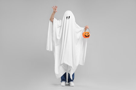 Woman in white ghost costume holding pumpkin bucket on light gray background. Halloween celebrationの写真素材
