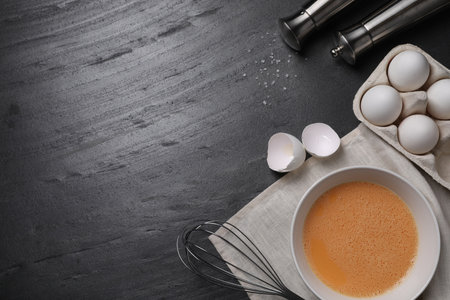 Metal whisk, beaten eggs and shells on dark gray background, flat lay. Space for text