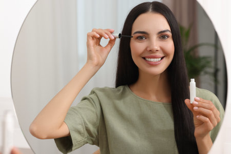 Beautiful woman applying serum onto her eyelashes in room, reflection in mirror. Cosmetic productの写真素材