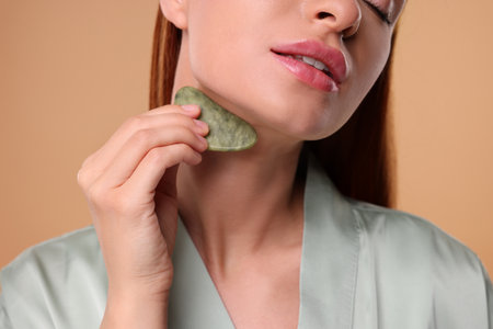 Young woman massaging her face with jade gua sha tool on pale orange background, closeupの写真素材