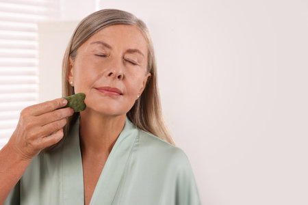 Woman massaging her face with jade gua sha tool in the bathroom. Space for textの写真素材