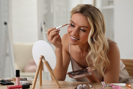 Beautiful makeup. Smiling woman applying eyeshadows in front of mirror indoorsの写真素材