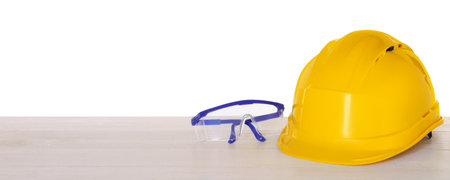 Hard hat and goggles on light wooden table against white background. Safety equipment