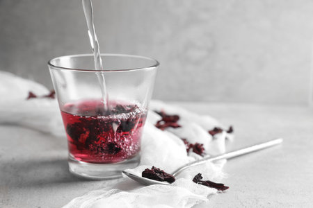 Making aromatic hibiscus tea. Pouring water into glass with dried roselle calyces and spoon on light table, space for textの写真素材