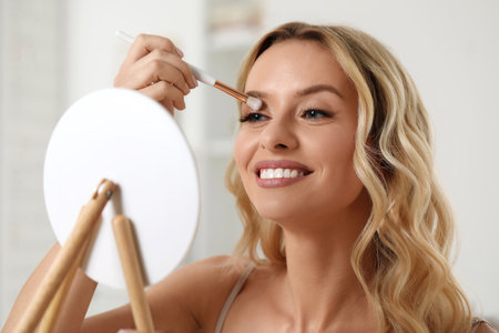 Beautiful makeup. Smiling woman applying eyeshadows in front of mirror indoorsの写真素材