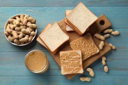 Delicious toasts with peanut butter and nuts on light blue wooden table, flat lay