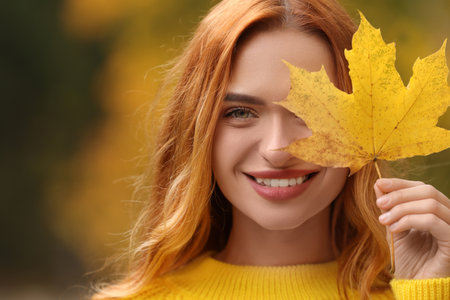 Smiling woman covering eye with autumn leaf outdoors, closeup. Space for textの写真素材