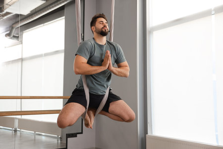 Young man meditating in fly yoga hammock indoors