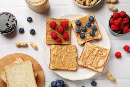 Delicious toasts with peanut butter, raspberries, blueberries and nuts on white tiled table, flat layの写真素材