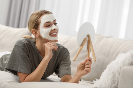 Young woman with face mask looking into mirror on sofa at home. Spa treatmentsの写真素材