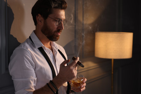 Handsome man with glass of whiskey smoking cigar at home. Space for textの写真素材