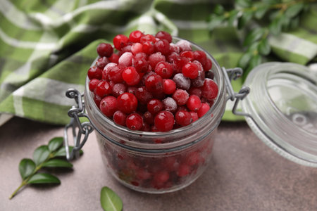 Frozen red cranberries in glass jar and green leaves on brown textured table, closeupの写真素材