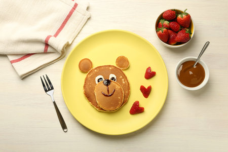 Creative serving for kids. Plate with strawberries, cute bear made of pancakes on light wooden table, flat layの写真素材