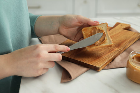 Woman spreading tasty nut butter onto toast at white table, closeup