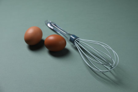 Whisk and raw eggs on pale blue background