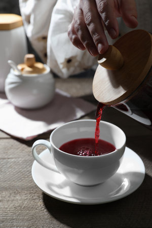 Woman pouring freshly brewed hibiscus tea from teapot into cup at wooden table, closeupの写真素材