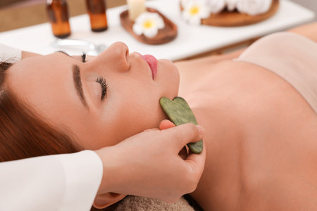 Young woman receiving facial massage with jade gua sha tool in beauty salon, closeupの写真素材