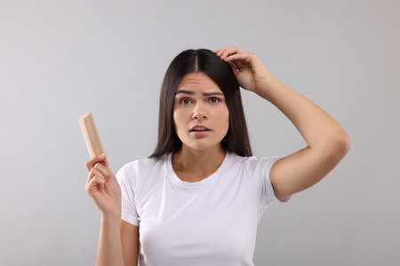 Emotional woman with comb examining her hair and scalp on gray backgroundの写真素材