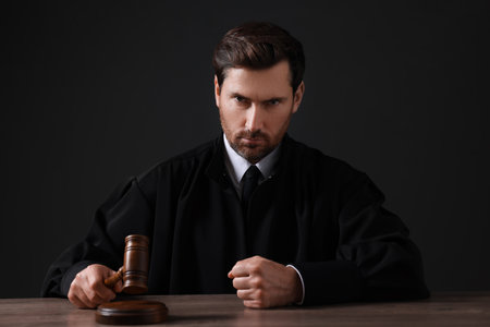 Judge with gavel sitting at wooden table against black backgroundの写真素材